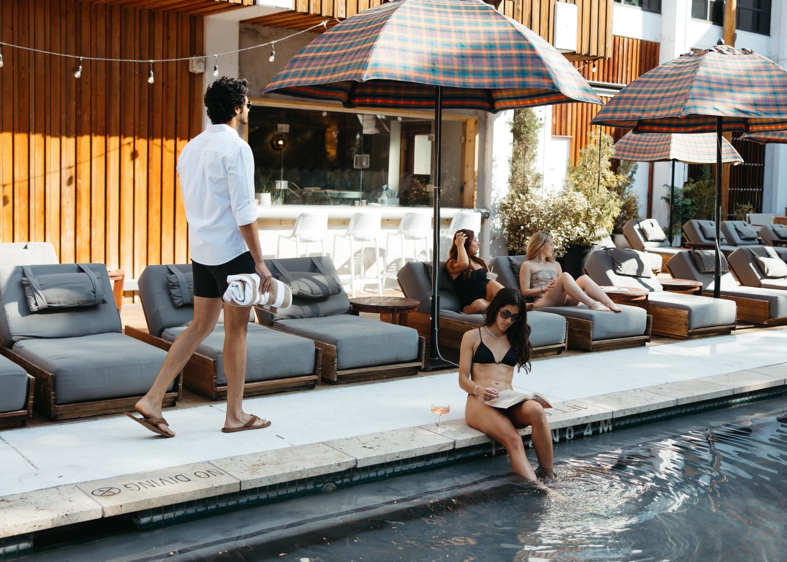 Man walking by a poolside lounge with women relaxing under umbrellas.