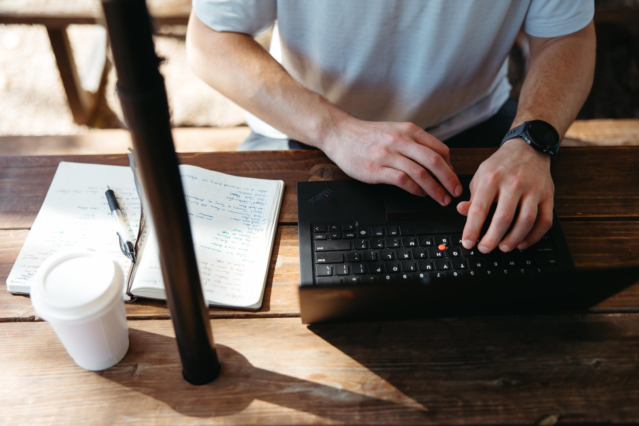 Close-up of a person typing on a laptop at a wooden table with a notebook and coffee nearby.