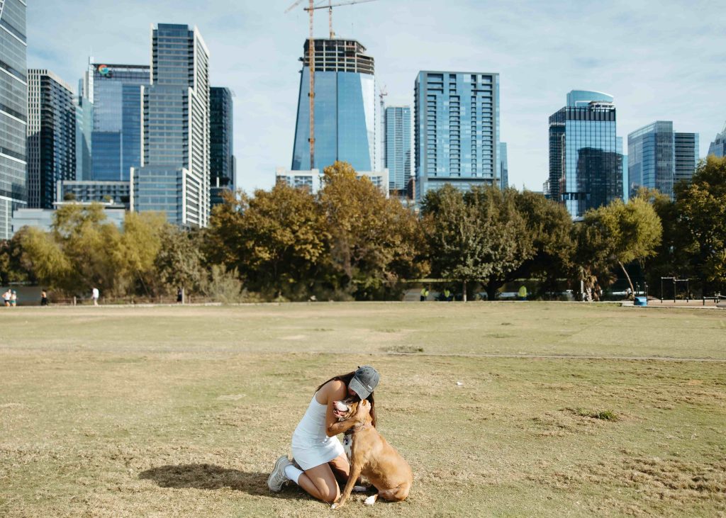 Woman kneeling in a grassy park, hugging her dog with Austin skyscrapers in the background.