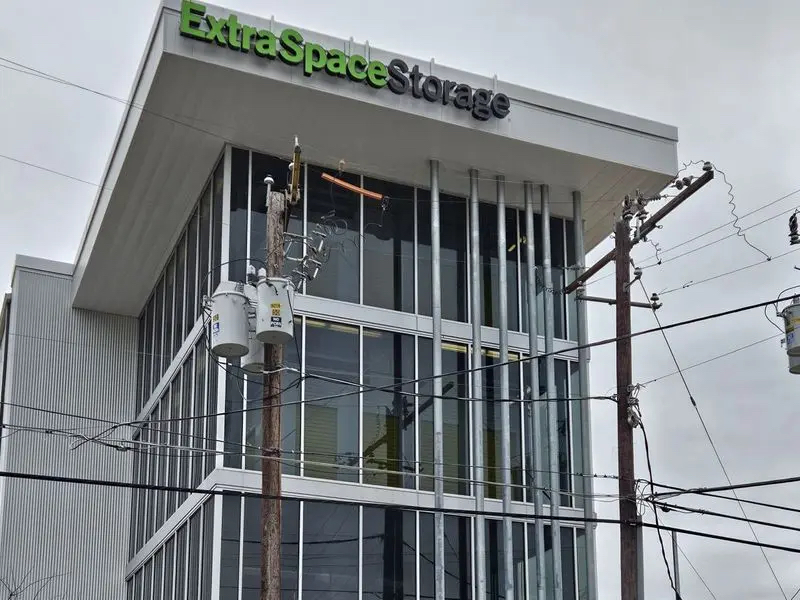 A modern Extra Space Storage building with large glass windows, white vertical panels, and green signage, seen from below with utility poles and power lines in front, under a cloudy sky.