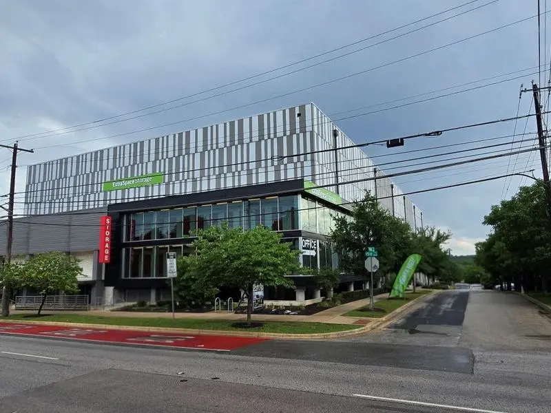 A modern, multi-story building with vertical gray and white stripes, featuring a green Enterprise sign and a red Storage sign for Austin self storage. The street is empty, lined with trees, and the sky above is cloudy.