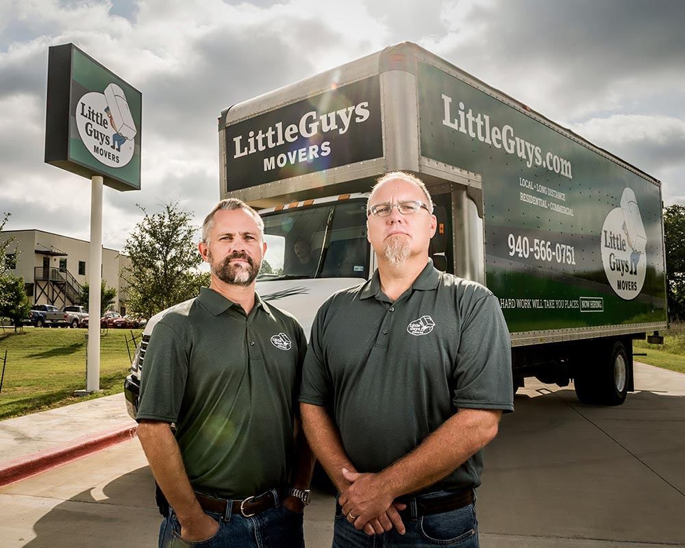 Two men in matching polo shirts stand in front of a Little Guys Movers box truck parked near a sign for the same company on a sunny day.