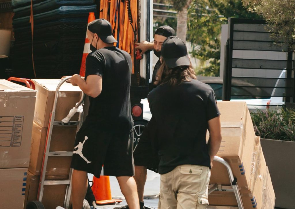Three movers from Pack N’ Go, one of the best Austin moving companies, wearing casual clothes and caps are moving large cardboard boxes with hand trucks from a moving truck parked outdoors on a sunny day.
