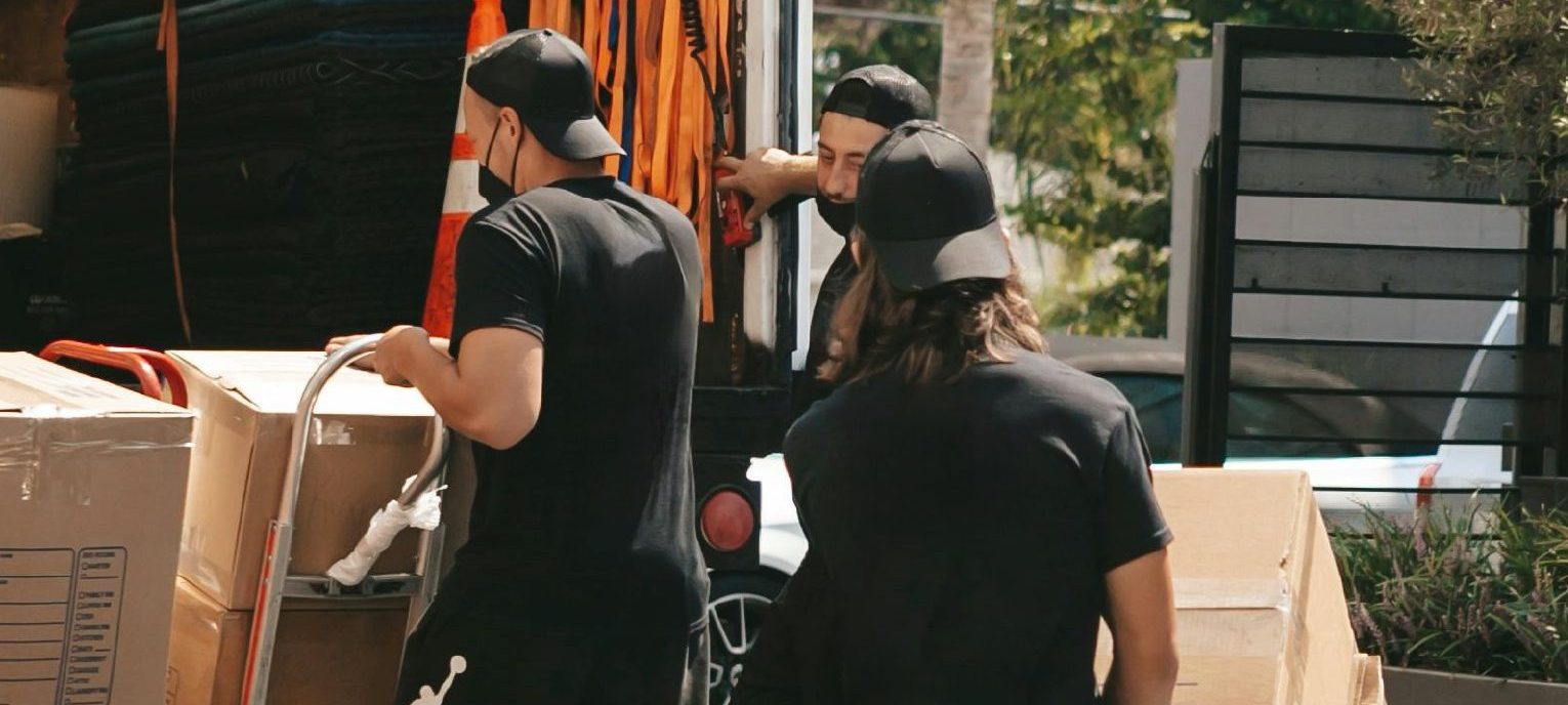 Three movers from Pack N’ Go, one of the best Austin moving companies, wearing casual clothes and caps are moving large cardboard boxes with hand trucks from a moving truck parked outdoors on a sunny day.