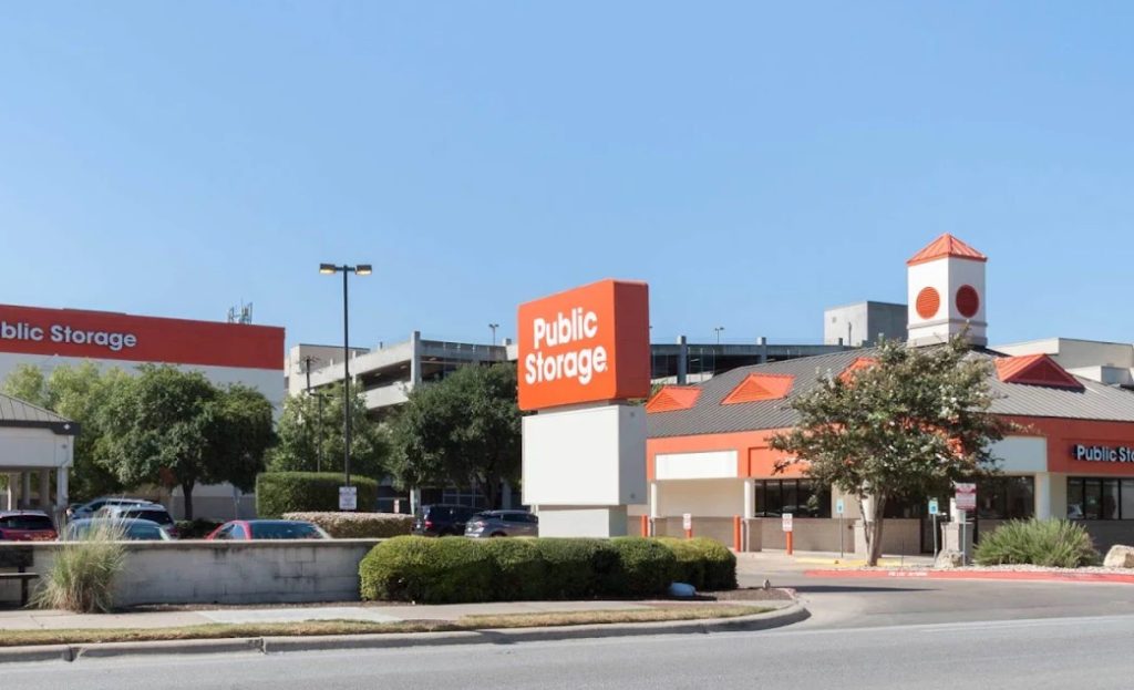 A Public Storage facility with orange and white signage and buildings is pictured on a sunny day, offering convenient Austin self storage, with a multi-level parking structure and several trees in the background.