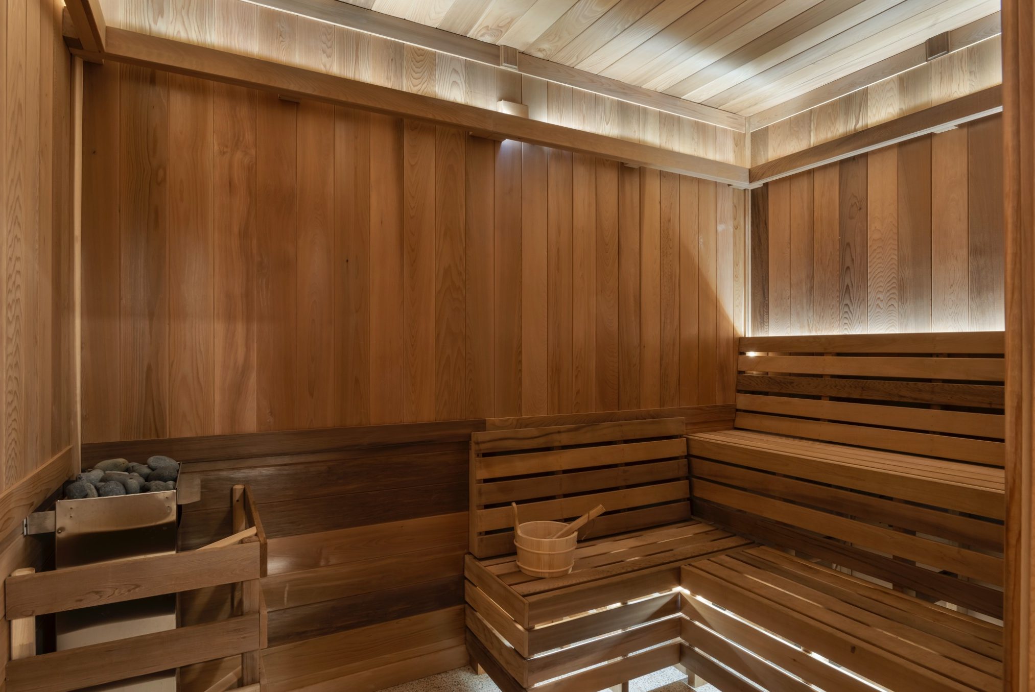 A modern paseo sauna with wooden benches, a bucket and ladle on the seat, and a rock-filled heater in the corner. Warm light shines through slats in the ceiling and upper walls, creating an inviting atmosphere.