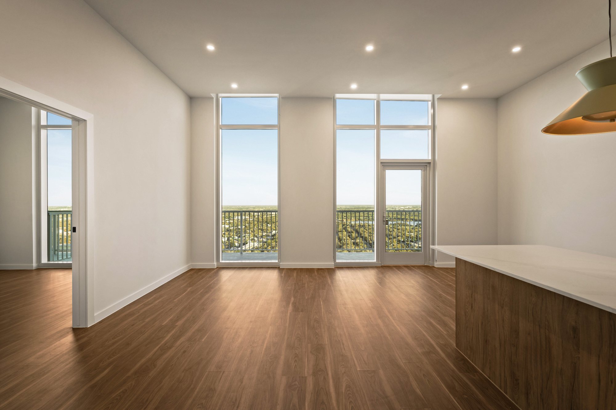 Spacious, modern empty room with wood floors, tall windows, and a door opening to a balcony with sweeping Paseo views of trees and sky; part of a kitchen island and a hanging light are visible on the right.