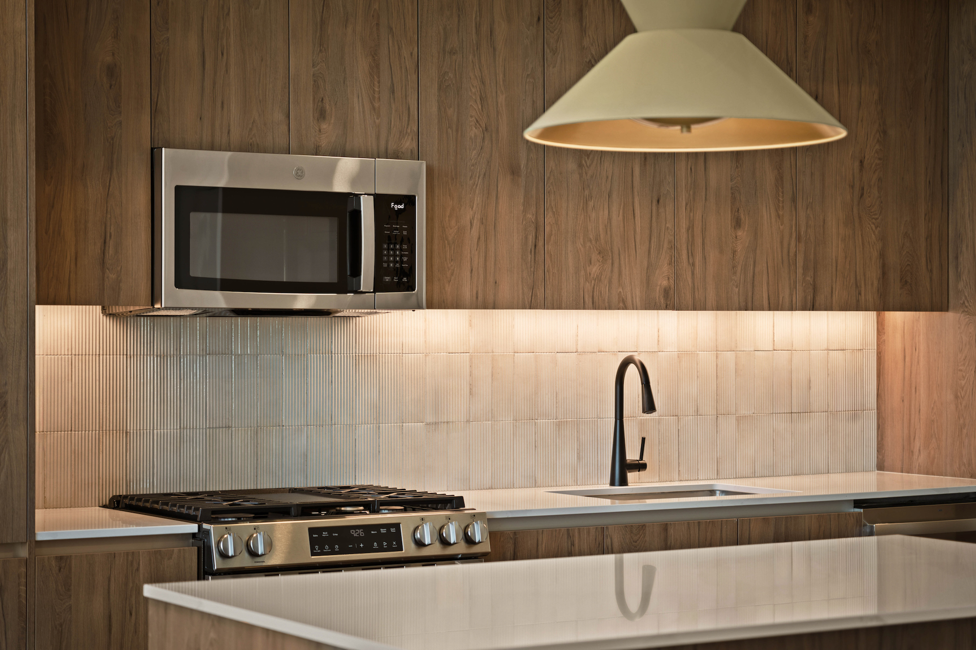 Modern Paseo kitchen with wood-paneled cabinets, a stainless steel microwave above a gas stove, black faucet by a white sink, tile backsplash, and a large, cream-colored pendant light overhead.