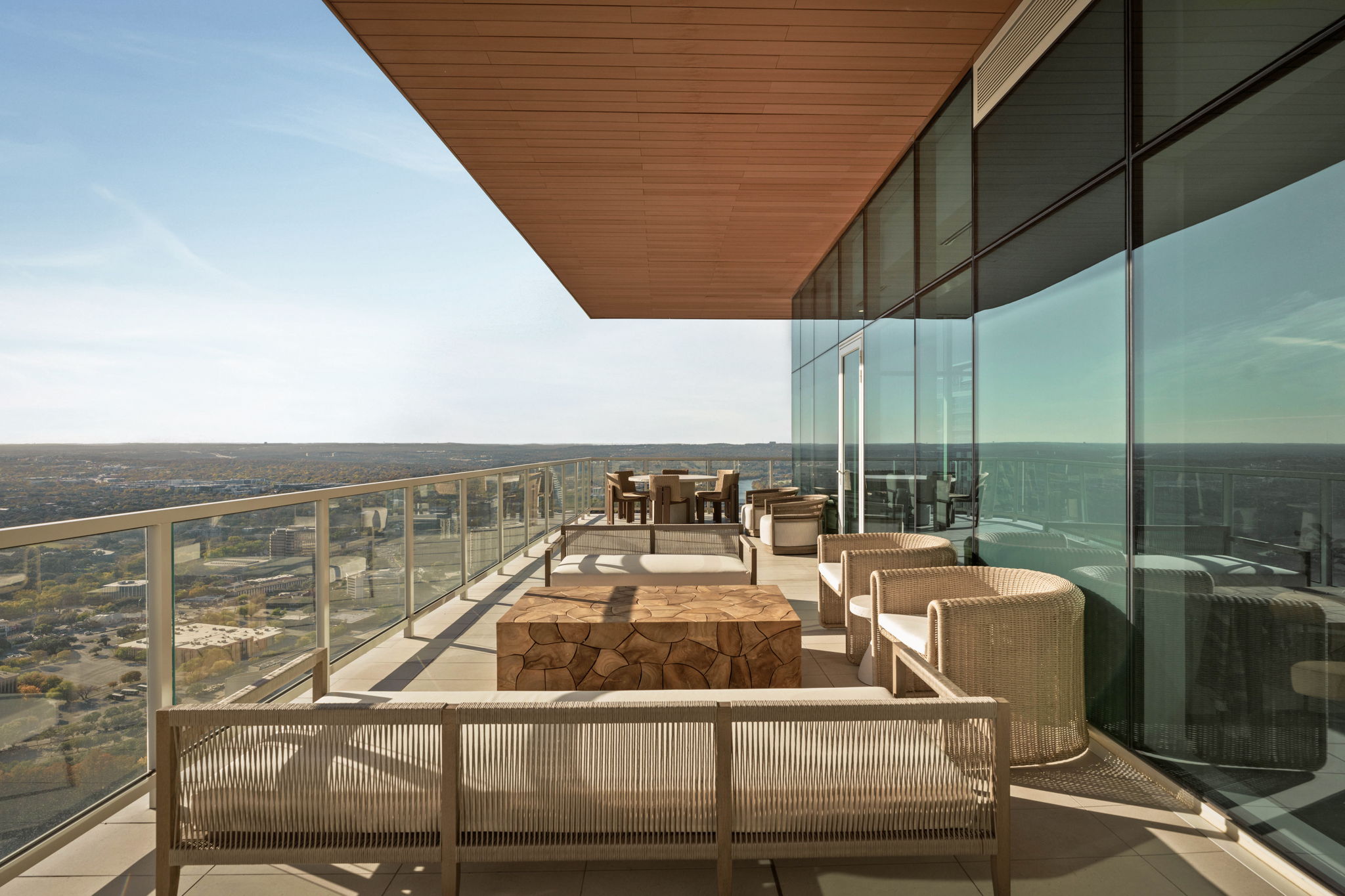 A modern high-rise balcony, reminiscent of luxury penthouses, with glass railings and large windows, features woven outdoor furniture and wooden tables overlooking a wide cityscape under a clear sky.