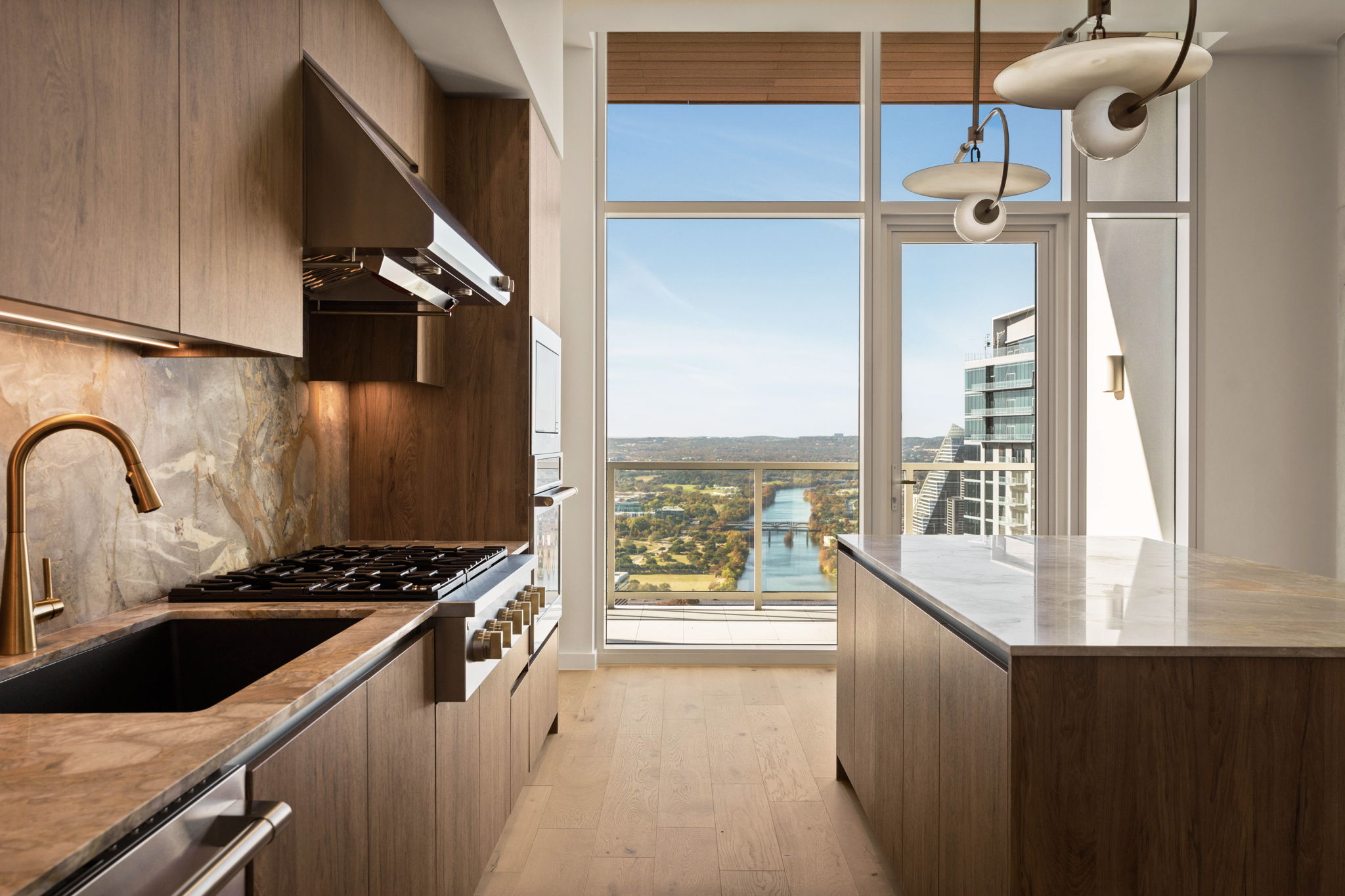 Modern kitchen with wood cabinets, marble countertops, gold faucet, and large windows offering stunning penthouse city and river views. Pendant lights hang from the ceiling as natural light fills the space.