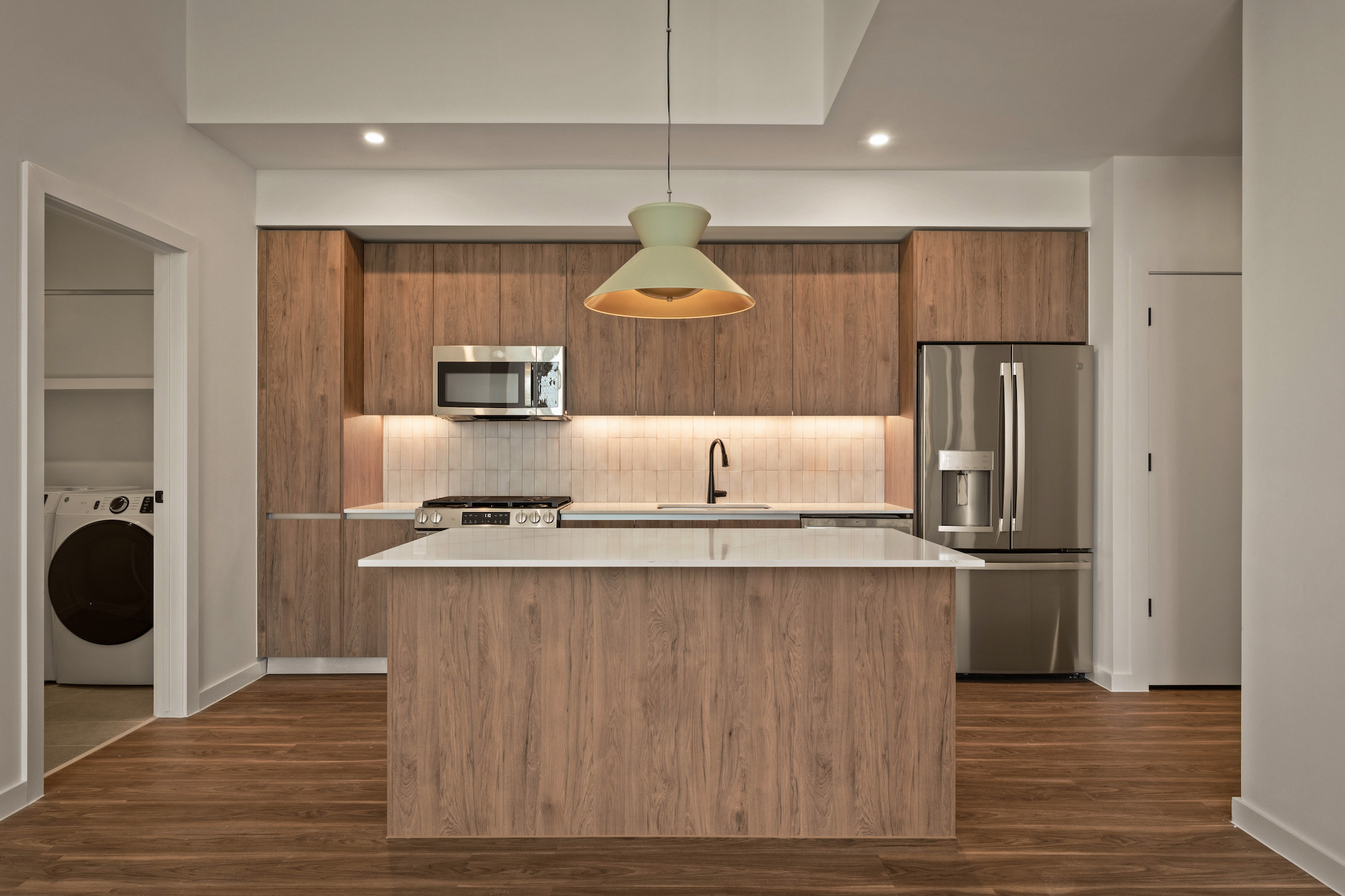 Modern kitchen with wood cabinets, a white island countertop, stainless steel appliances including a microwave and fridge, a stylish light fixture above the island, and laundry area visible through an open doorway in your Paseo home.