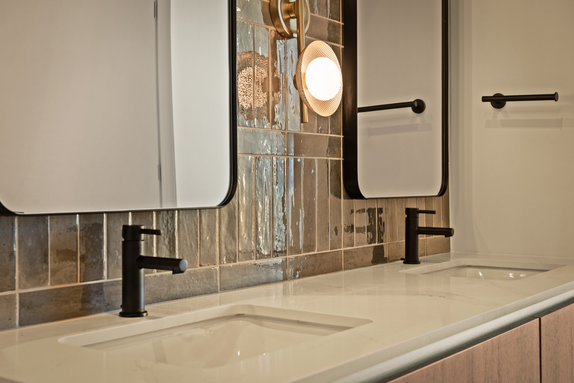 Modern bathroom with two rectangular sinks, black faucets, and matching towel racks. Above the sinks are rectangular mirrors and a wall sconce with a round bulb, all set against glossy brown and beige tile backsplash for a Paseo-inspired touch.
