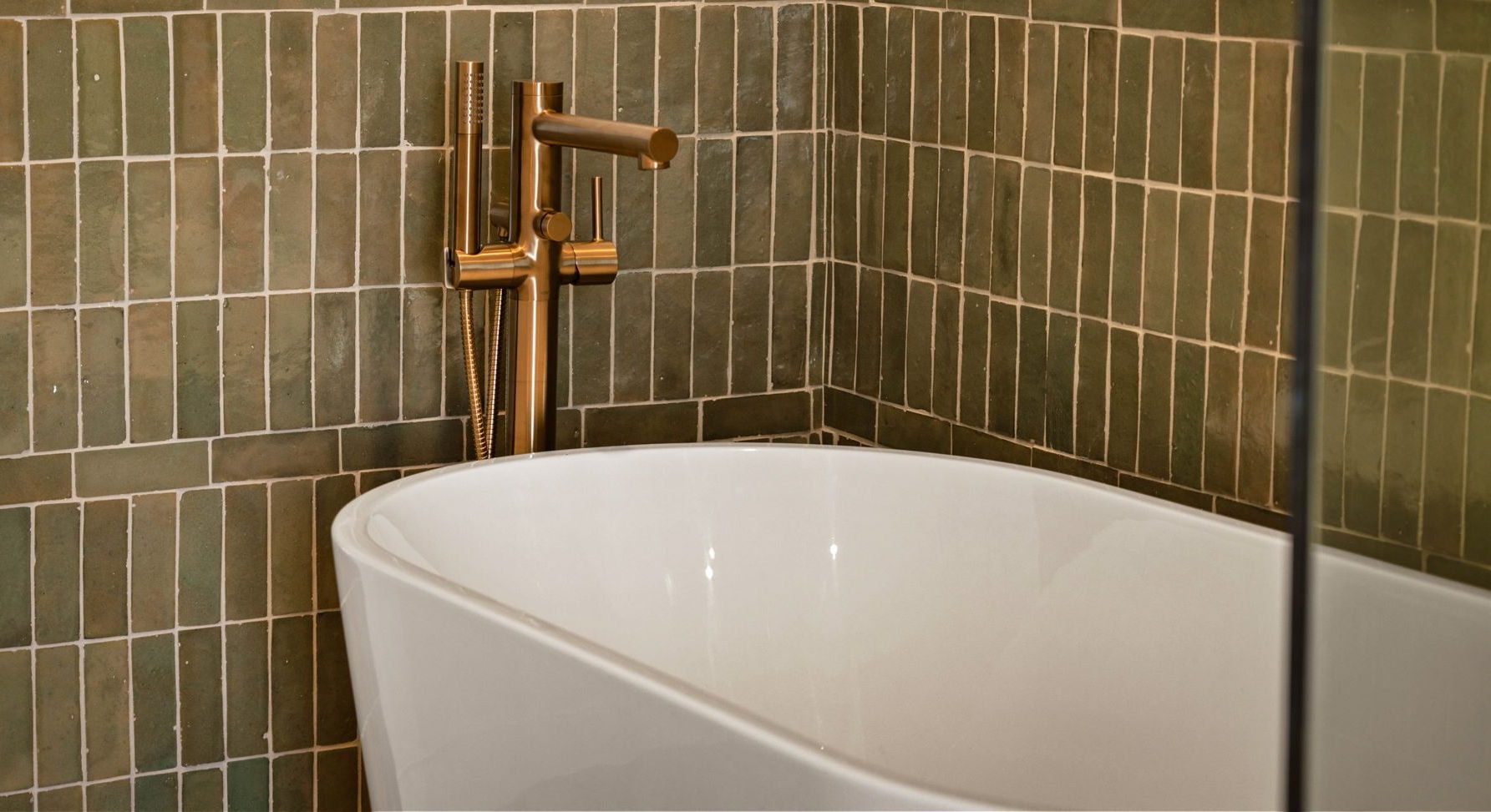 A modern bathroom, reminiscent of upscale penthouses, features a freestanding white bathtub with a gold faucet set against a wall of green rectangular tiles arranged in a grid pattern.