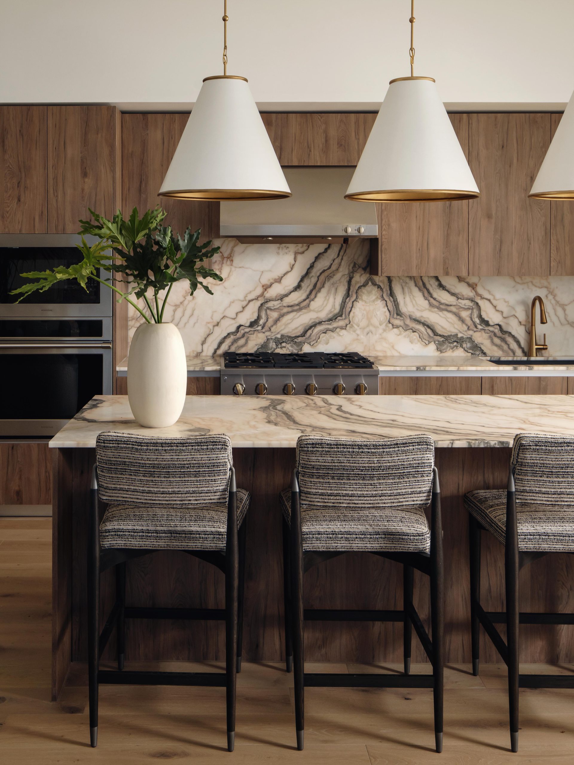 A modern kitchen, perfect for penthouses, featuring a marble island with three gray upholstered barstools, a vase with green leaves, wood cabinetry, a marble backsplash, a stove, and three large white pendant lights.