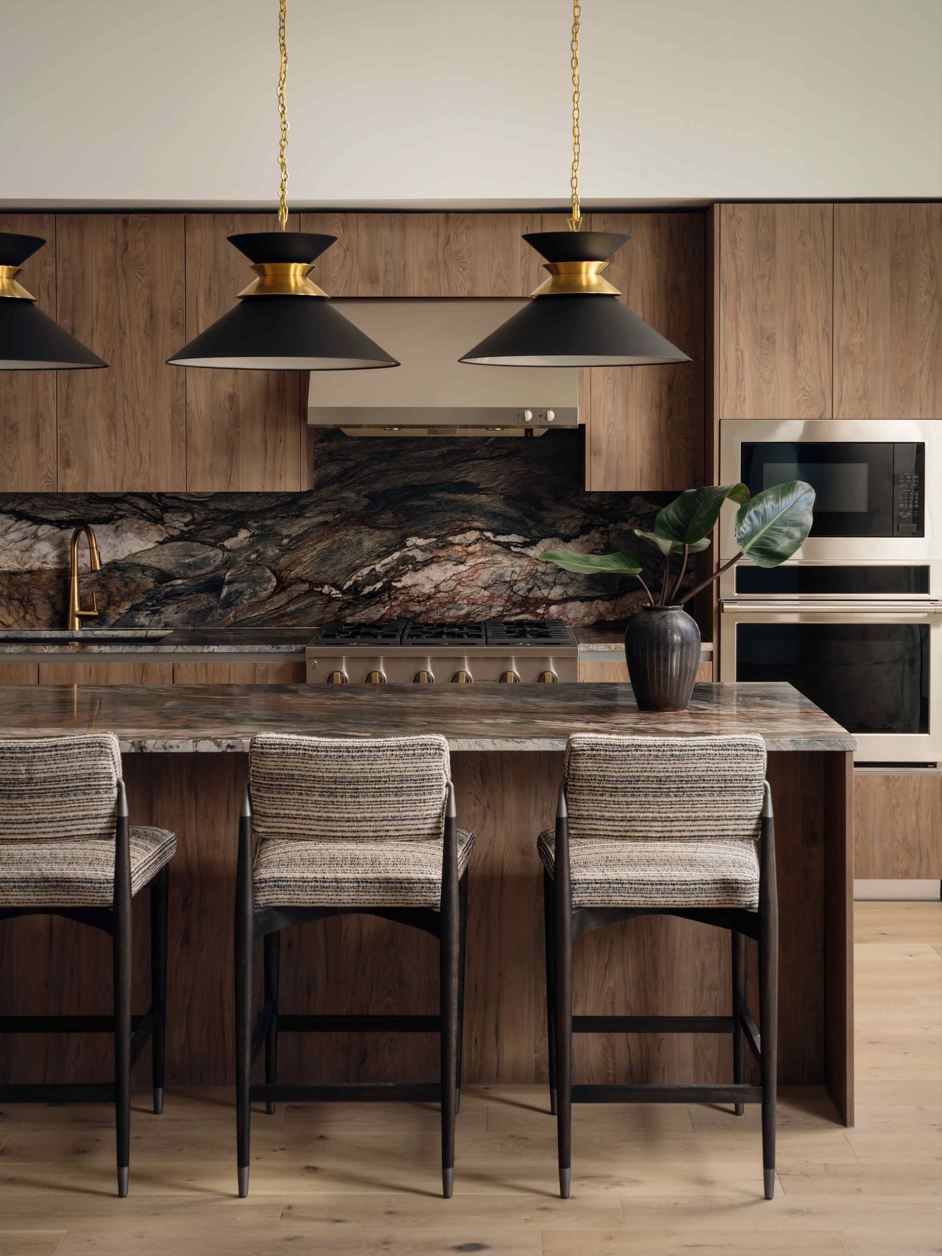 Modern kitchen with wood cabinetry, a marble-patterned backsplash, and an island with three upholstered bar stools—perfect for penthouses. Black and gold pendant lights hang above, while built-in ovens and a plant in a vase complete the elegant space.