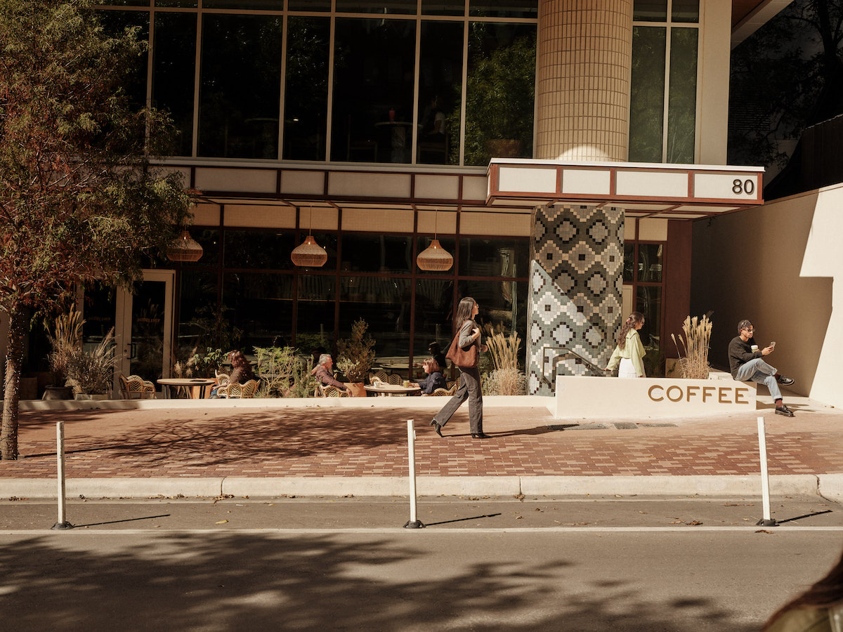 People sit and relax outside a modern coffee shop with large windows and geometric tile décor, nestled beneath luxury apartments Austin TX. A woman walks past on the sidewalk, while another person sits near a sign that says 