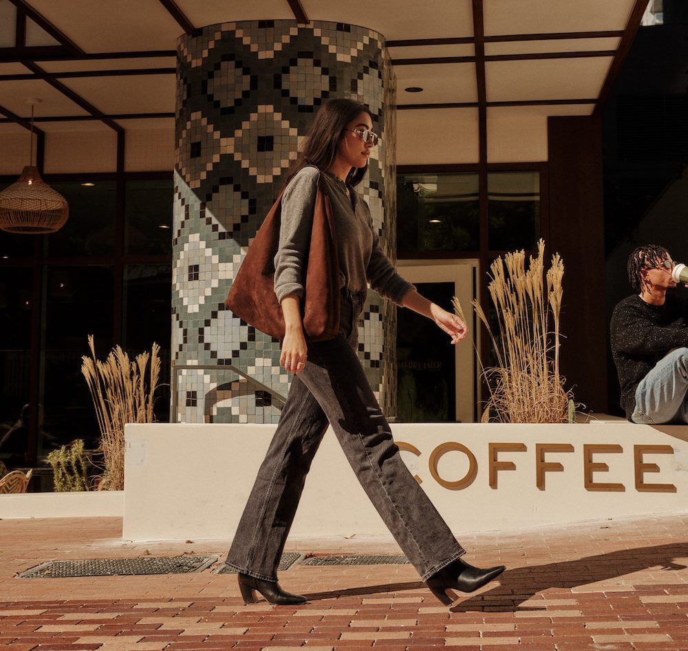 A woman with long dark hair, sunglasses, and a brown bag walks past a coffee shop with geometric tile patterns on the wall, just steps from downtown Austin apartments, while a person sits nearby drinking from a cup.
