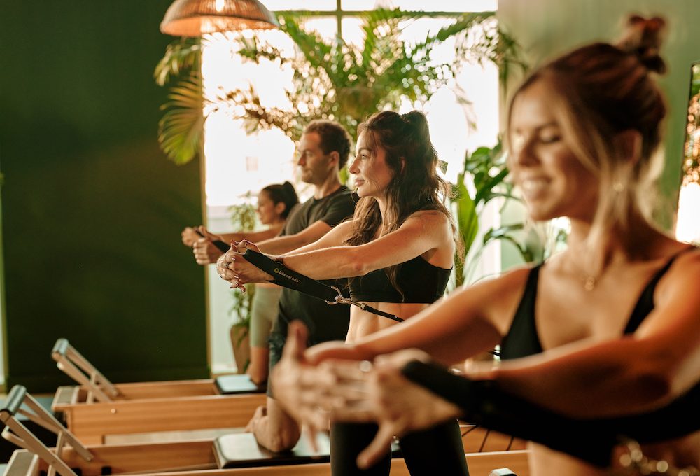 Four people exercising on Pilates reformer machines in a well-lit, green-walled studio with plants and hanging lights. They are focused and wearing athletic clothing, performing arm stretches facing forward.