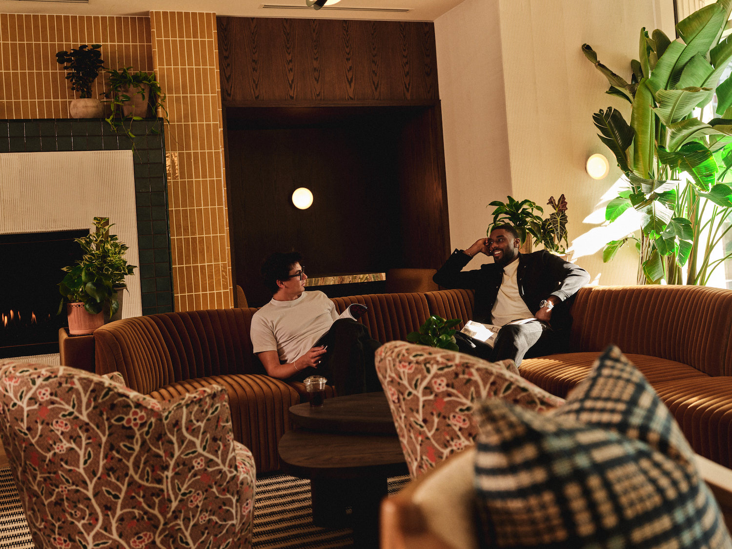 Two men sit and talk on a brown curved sofa in a stylish, modern lounge at luxury apartments in Austin, TX, surrounded by plants, patterned chairs, a fireplace, and warm lighting.