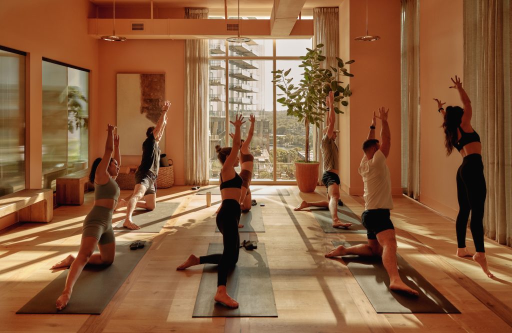A group of people practice yoga in a sunlit studio with large windows, wooden floors, and potted plants—an amenity commonly found in luxury apartments Austin TX residents enjoy. They kneel on mats with arms raised overhead, facing the front.