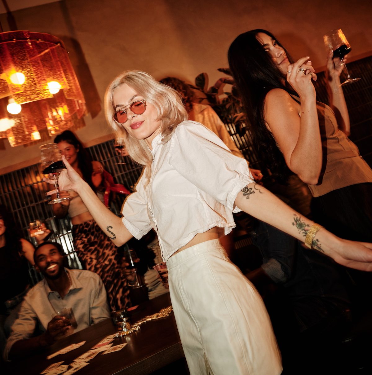 A woman with blonde hair and sunglasses dances while holding a glass of wine at a lively indoor party in one of the stylish downtown Austin apartments. Others in the background are smiling, chatting, and enjoying drinks in the warm, festive atmosphere.