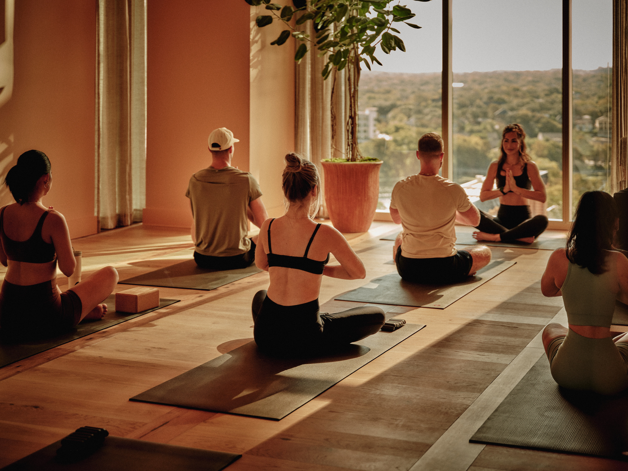 A group of people practice yoga, seated cross-legged on mats in a sunlit studio with large windows and a potted plant, while an instructor leads the session—an amenity often found at luxury apartments Austin TX residents love.