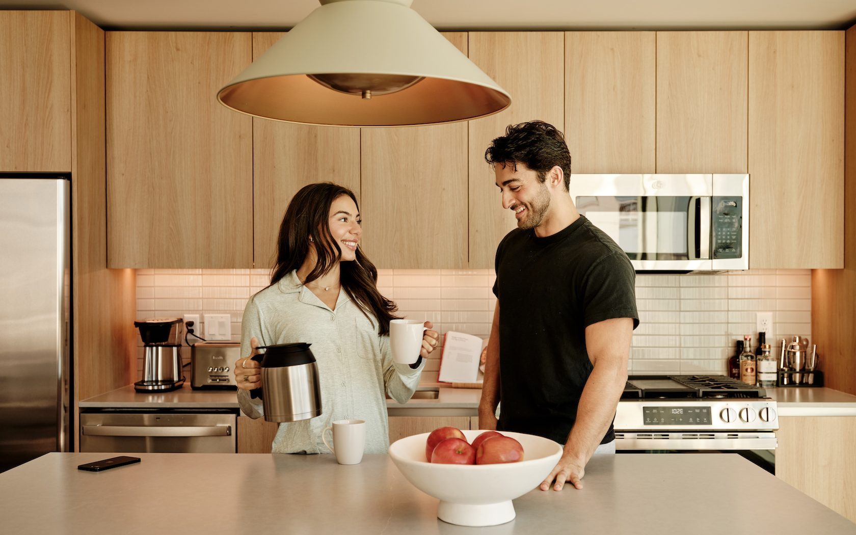 A smiling woman and man stand in a modern kitchen of luxury apartments Austin TX. The woman holds a coffeepot and a mug, while the man leans on the counter next to a bowl of apples as they enjoy a pleasant conversation.
