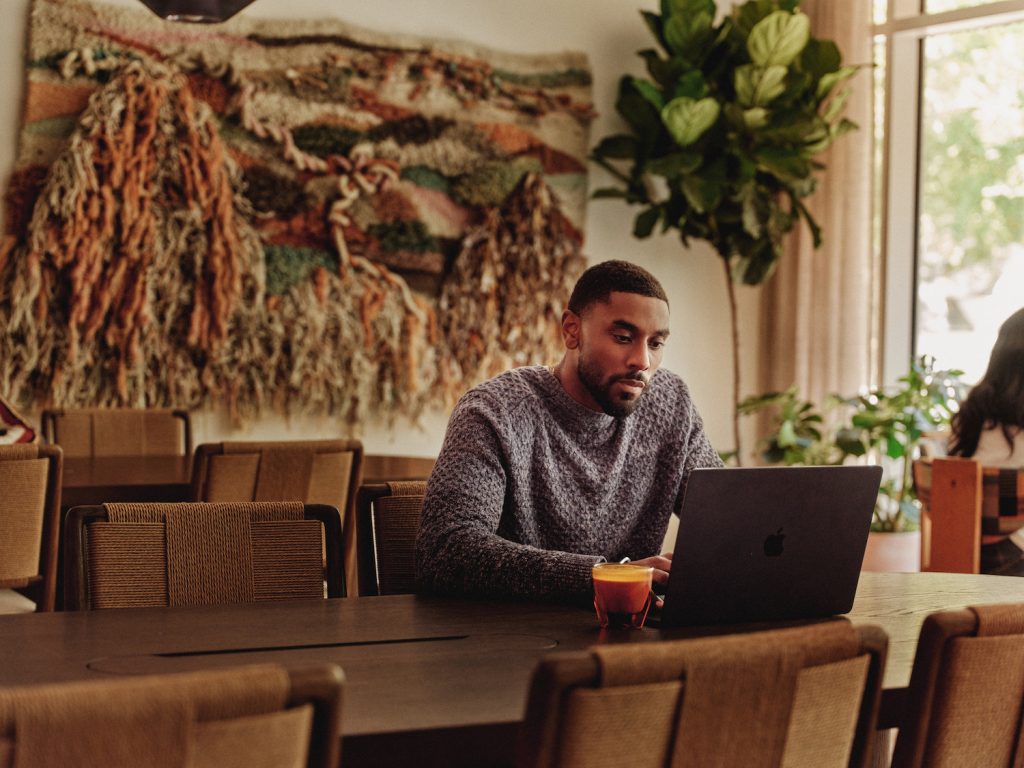 A man sits alone at a large table in luxury apartments Austin TX, working on a laptop with a drink beside him. Behind him is a decorative woven wall hanging and a large potted plant as sunlight streams through the window.