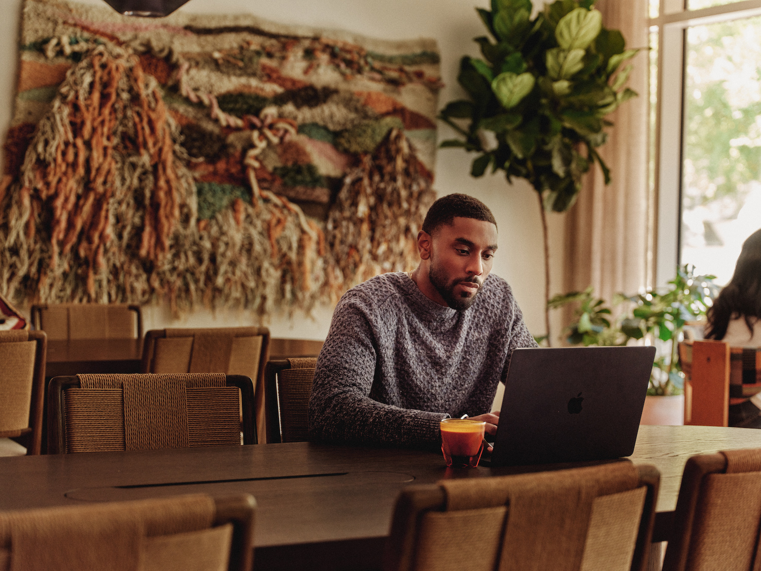 A man sits alone at a large table in luxury apartments Austin TX, working on a laptop with a drink beside him. Behind him is a decorative woven wall hanging and a large potted plant as sunlight streams through the window.
