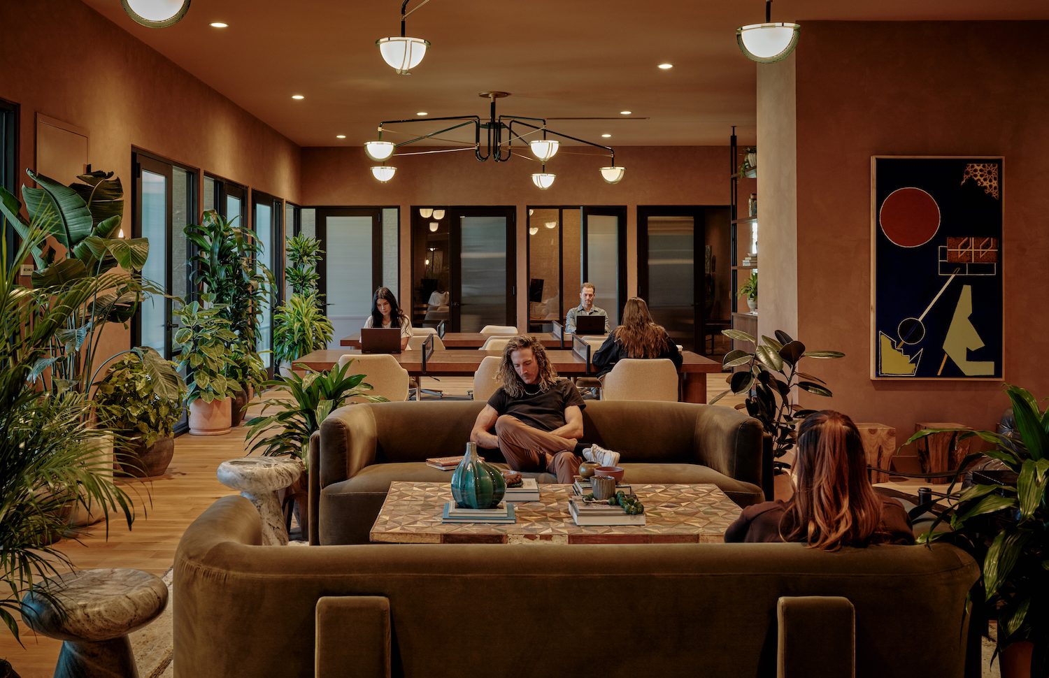 A cozy, modern lounge in luxury apartments Austin TX, with people working and relaxing on sofas, surrounded by indoor plants, warm lighting, and contemporary wall art. Large windows bring in natural light.