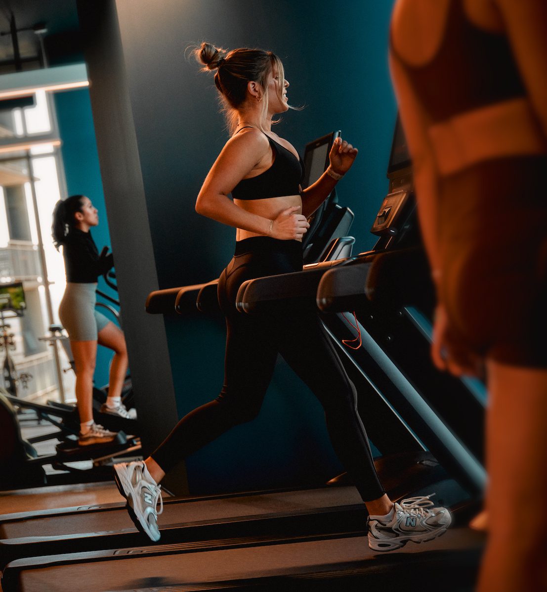 Three women focus on wellness as they exercise indoors; one runs on a treadmill, another pedals a stationary bike, and a third stands nearby, all dressed in athletic wear within a modern gym.