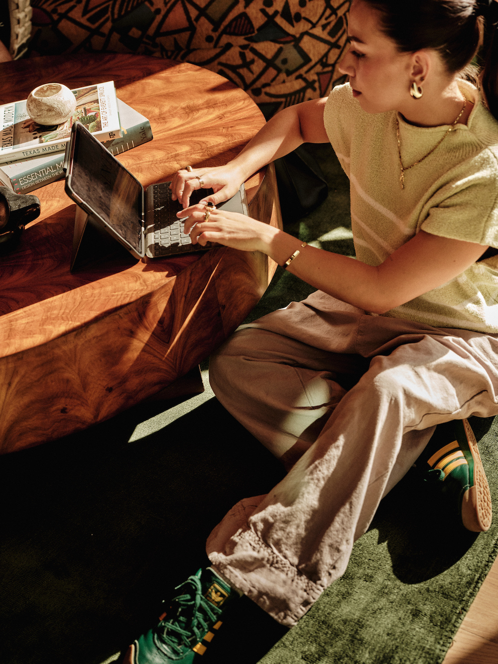 A person sits on the floor by a wooden coffee table in one of the luxury apartments Austin TX offers, typing on a laptop. They wear light-colored pants, a short-sleeved top, sneakers, and gold jewelry. Books and sunlight complete the cozy scene.
