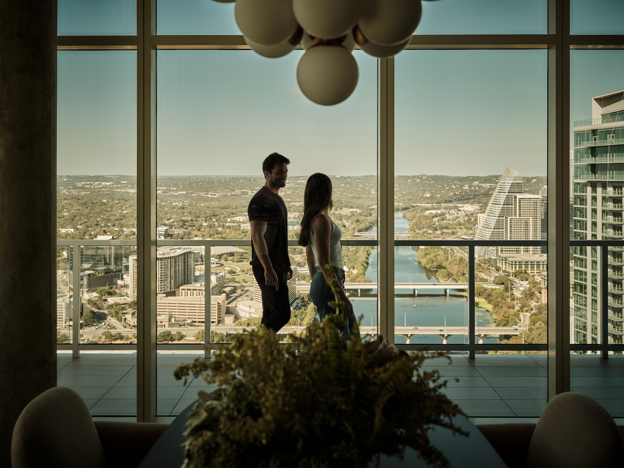 A man and woman stand facing each other by large floor-to-ceiling windows in one of the city's most luxurious penthouses, overlooking a river, tall buildings, and hills. A modern chandelier and dining table with flowers are in the foreground.