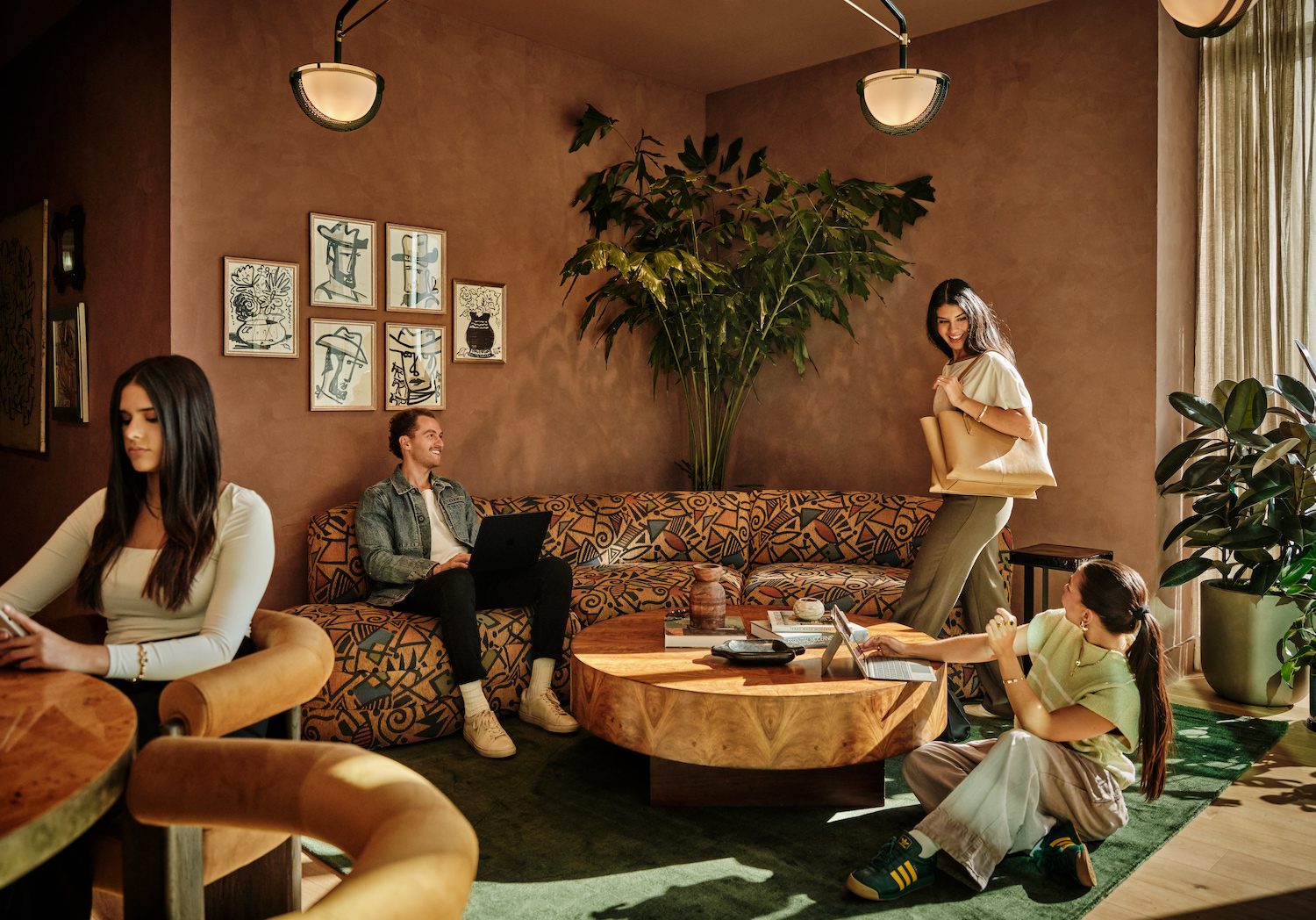 Four people work and interact in a cozy, modern lounge with warm earth tones and patterned furniture—just one of the inviting spaces found in luxury apartments Austin TX residents love. Large plants and framed art add to the welcoming vibe around a wooden table.