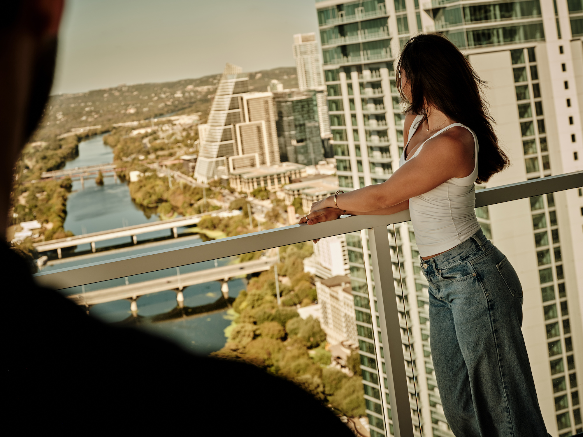 A woman in a white tank top and jeans stands on a high-rise balcony, gazing out over a river and cityscape with penthouses, modern buildings, and a bridge in the background. A person’s silhouette appears in the foreground.