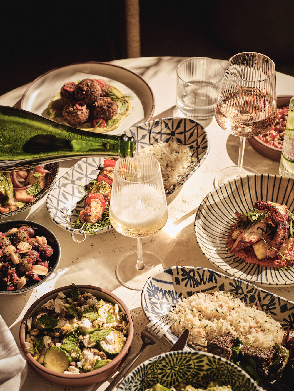 A table set with patterned bowls and plates of assorted Mediterranean dishes, including grilled vegetables, salads, rice, and meat—just the kind of dining experience you’d enjoy in luxury apartments Austin TX, as sunlight spills over poured wine.