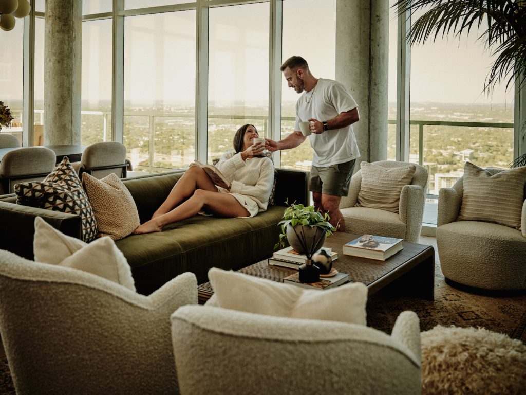 A man hands a drink to a woman relaxing on a green sofa in a modern, sunlit penthouse living room with large windows, cozy chairs, a coffee table with books, and decorative cushions and plants.
