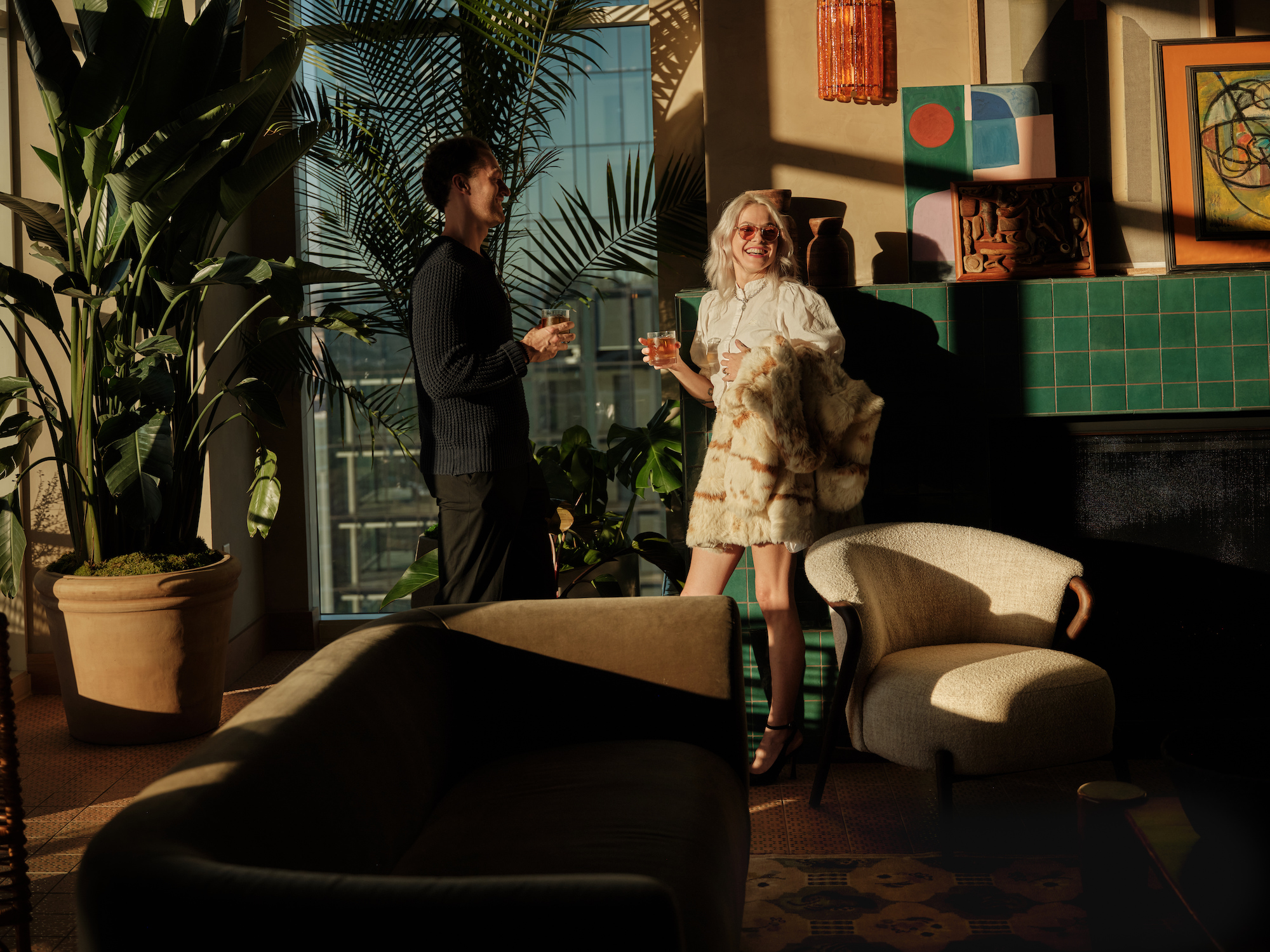 Two people are standing and holding drinks in a stylish, sunlit room with modern decor, green plants, and abstract artwork—classic features of luxury apartments Austin TX offers. Warm sunlight creates a relaxed, inviting atmosphere throughout the space.