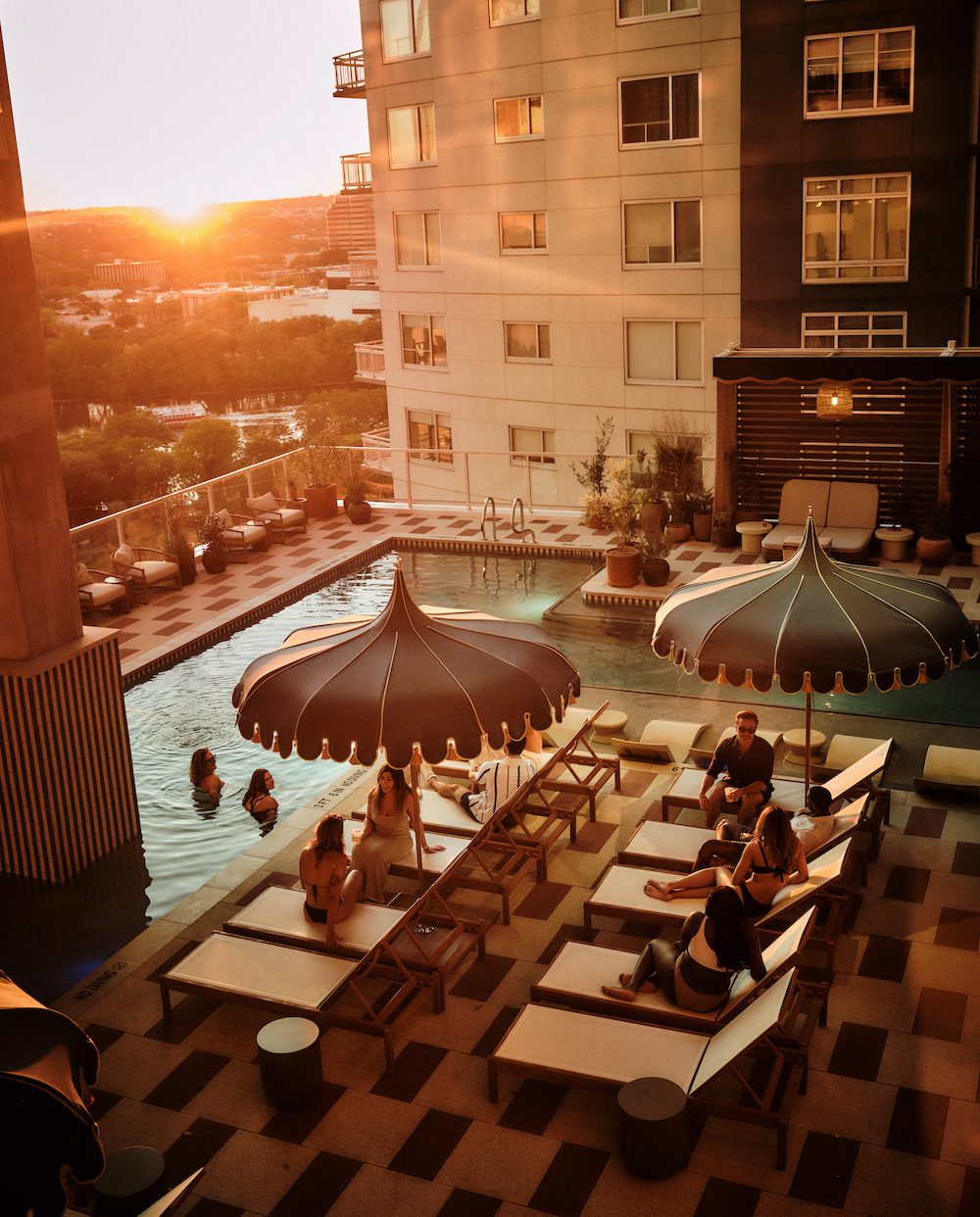 People relax on lounge chairs and swim in a rooftop pool at sunset at luxury apartments Austin TX, with large umbrellas providing shade, surrounded by a modern building and stunning city views in the background.