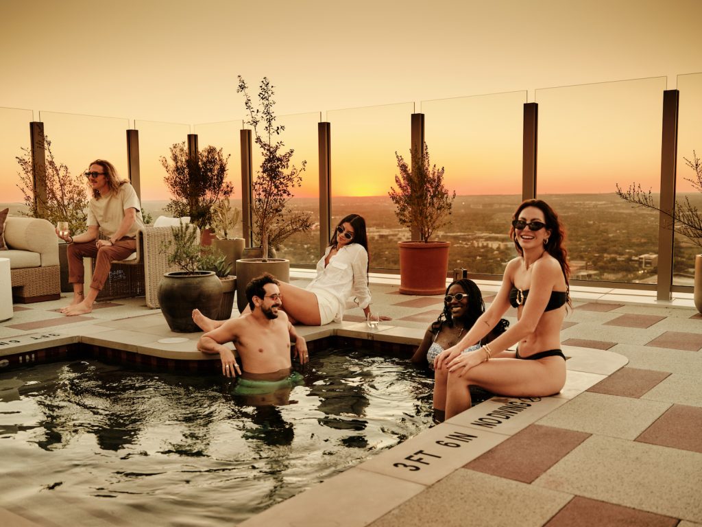 Five adults relax by a rooftop hot tub at sunset in luxury apartments Austin TX, chatting and smiling. Two women sit on the tub’s edge, a man and woman are in the water, and one man sits on a chair with cityscape and plants in the background.
