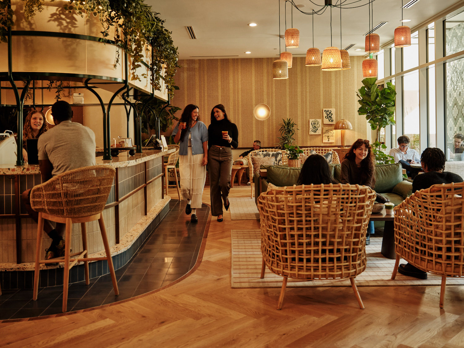 A modern, cozy café with people sitting on wicker chairs, chatting and working—much like the inviting community spaces found in luxury apartments Austin TX offers—while others relax or converse in a warmly lit, plant-filled setting.