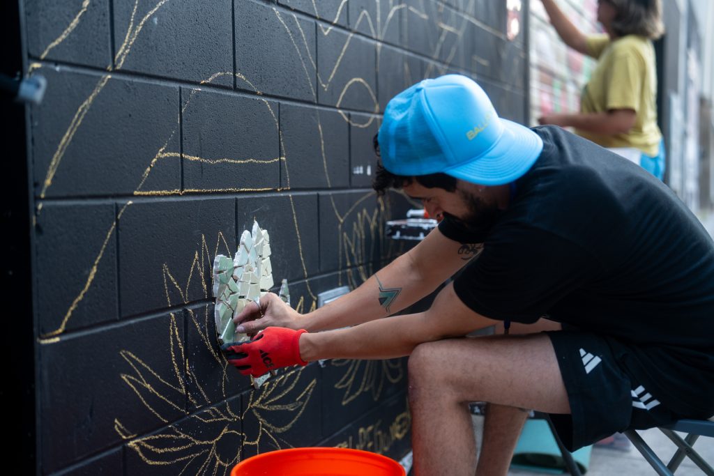 Artist Felipe Gomez placing hand-cut tile pieces onto a black brick wall during Mosaic Fest.