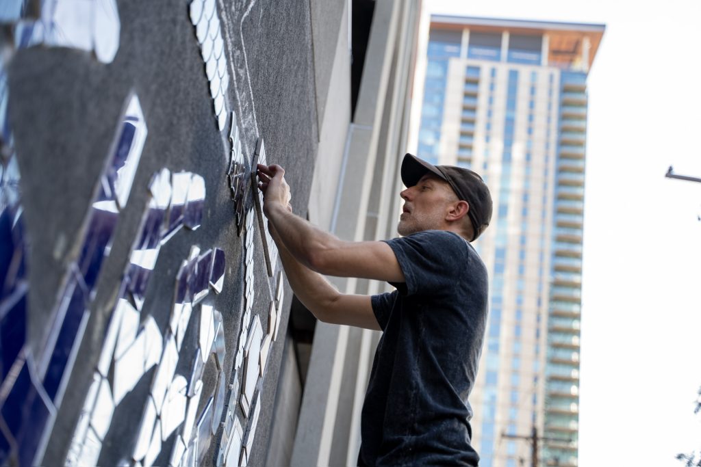 Mosaic artist Sleepisfamous installing reflective tiles onto a wall with Austin high-rises in the background during Mosaic Fest.