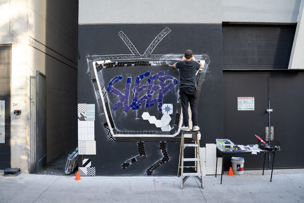Artist Sleepisfamous standing on a ladder installing a mosaic reading “SLEEP” on a black wall during Austin’s Mosaic Fest.