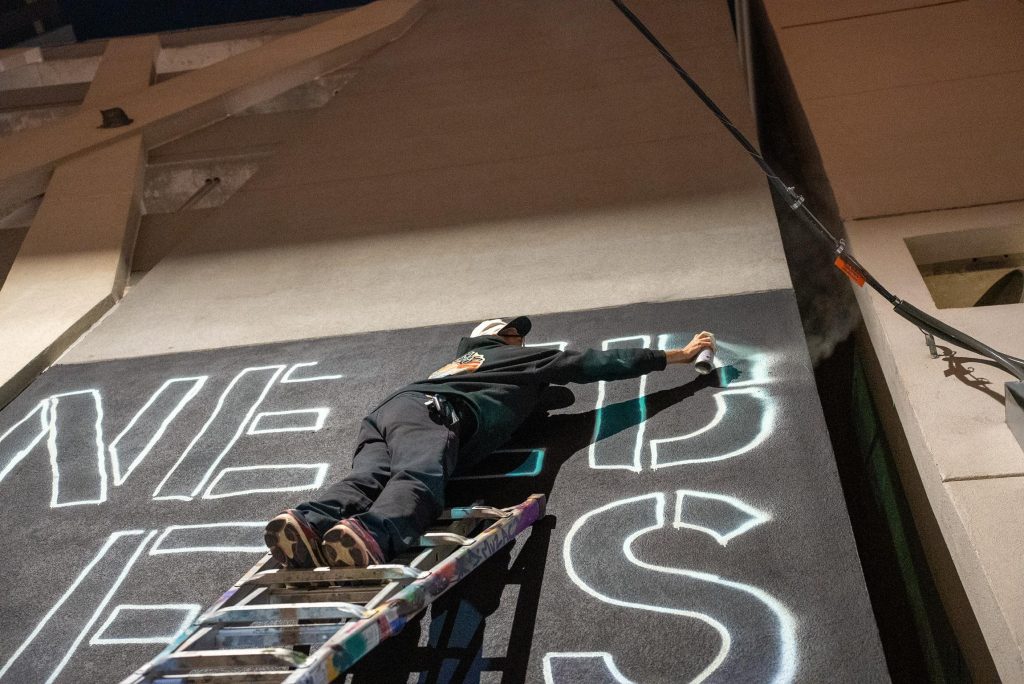 Artist J Muzacz spray-paints large outline letters on a tall black wall at night during Mosaic Fest in the Rainey Street alley.