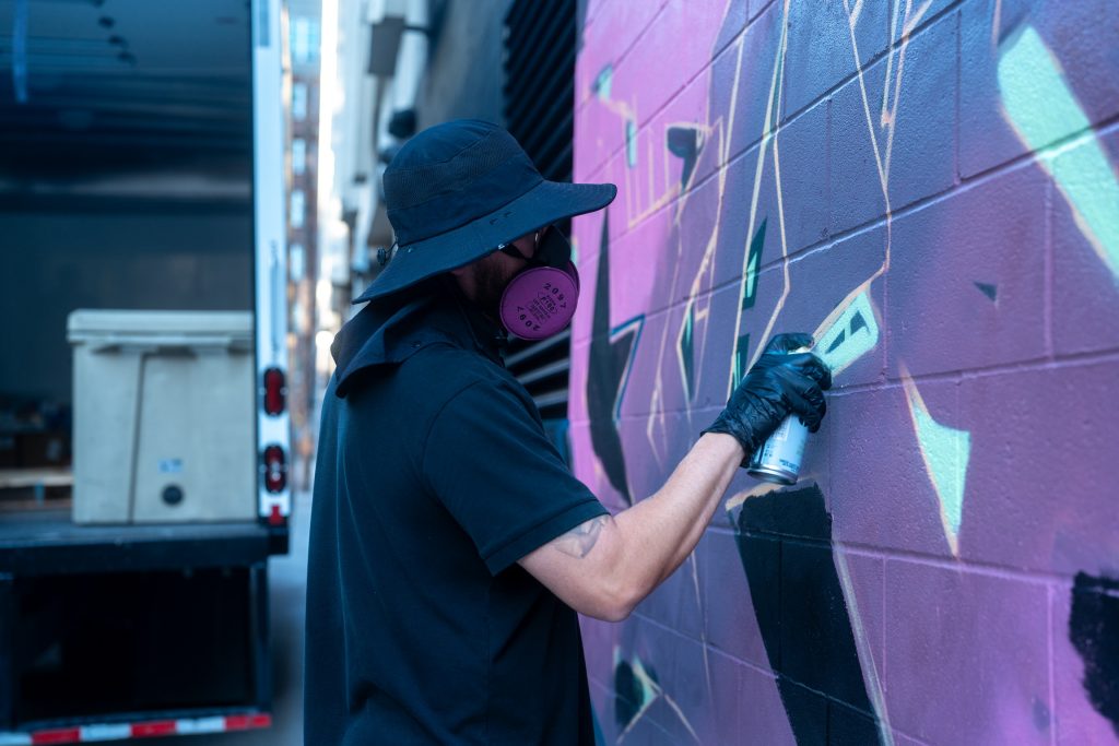 Spray-paint artist DFKTE wearing a mask and hat while painting a purple and teal mural on a brick wall during Mosaic Fest in Austin.