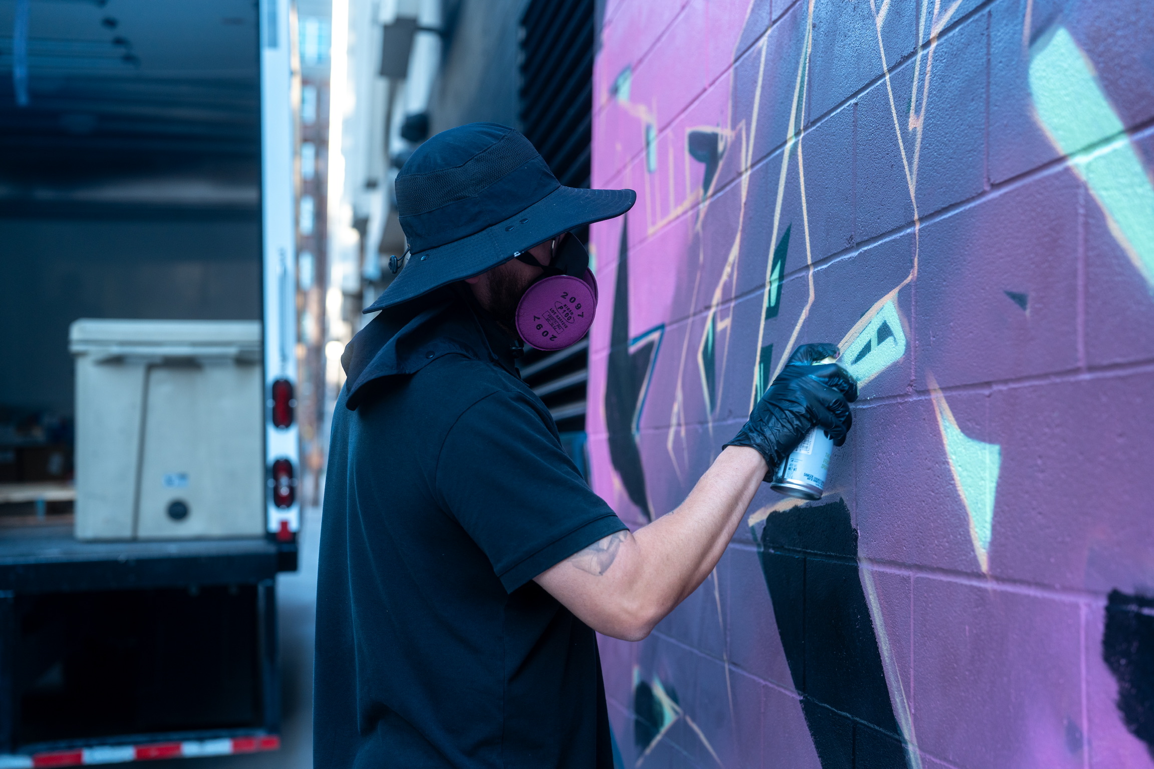 Spray-paint artist DFKTE wearing a mask and hat while painting a purple and teal mural on a brick wall during Mosaic Fest in Austin.