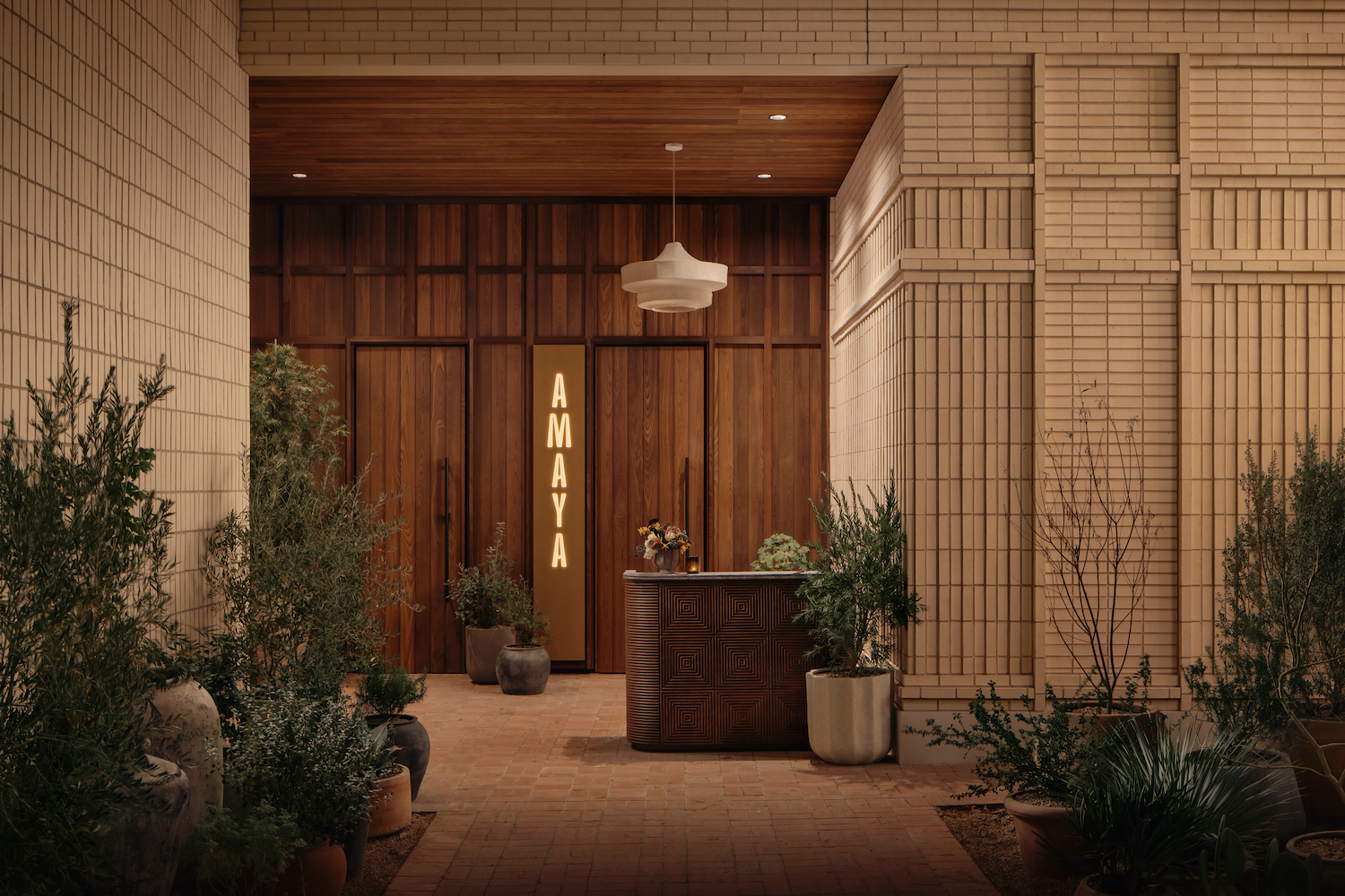 A warmly lit restaurant entrance with a wooden double door, a modern light fixture above, and a sign reading AMAYA welcomes guests along a brick paseo lined with potted plants and beige tiled walls.
