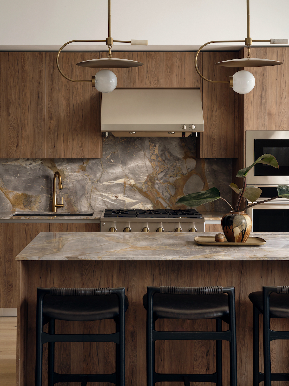 Modern Paseo kitchen with wood cabinets, marble backsplash and countertop, brass fixtures, two pendant lights, a gas stove, built-in oven, and three black stools at the island. A vase with leaves sits on the counter.