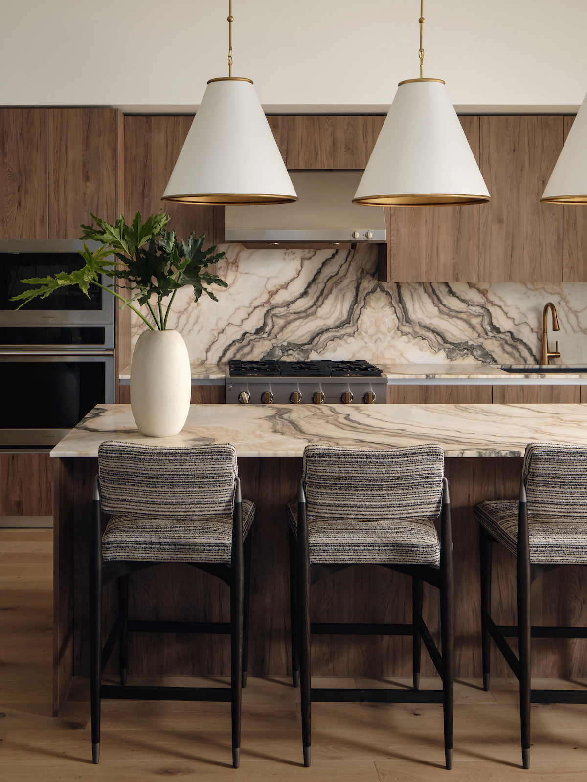 Modern Paseo kitchen with wood cabinets, a dramatic marble backsplash and countertop, three pendant lights, a vase with greenery, and three upholstered bar stools at the island.